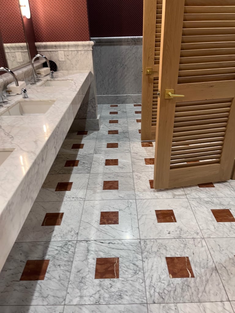 Luxury bathroom with marble and burgundy tile flooring, double vanity sink, wooden door, and gray marble backsplash