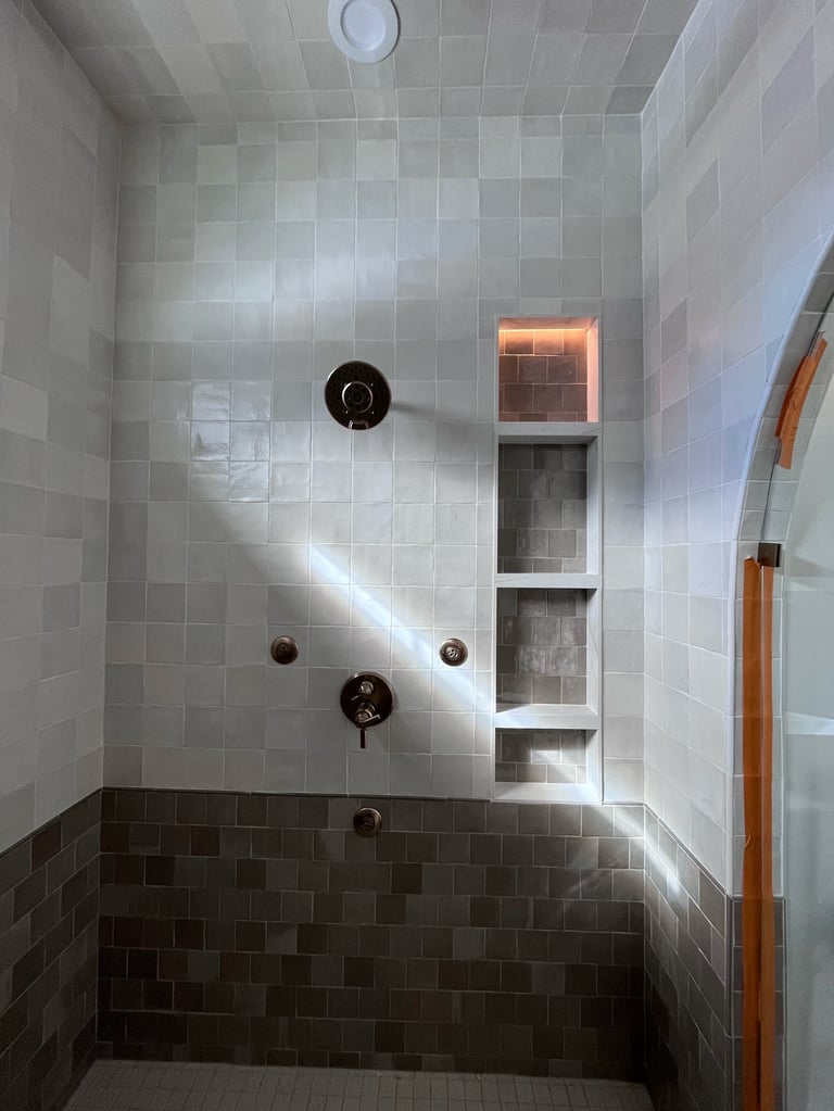 Modern shower stall with white and dark gray tile, bronze fixtures, built-in niches, and natural light from window
