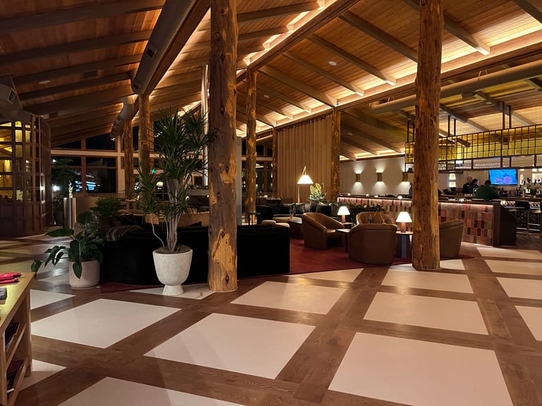 Modern hotel lobby with exposed wooden beams, geometric floor pattern, potted plants, and warm overhead lighting