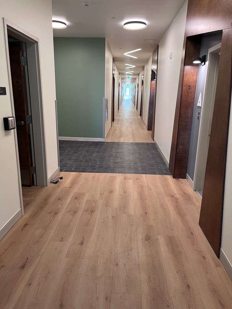 Modern office hallway with wood and tile flooring, multiple doors, recessed ceiling lights, and green accent wall