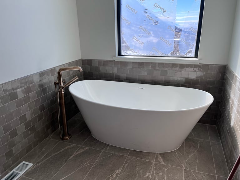 Modern white freestanding bathtub with bronze faucet in bathroom with gray tile walls and floor, window view in background