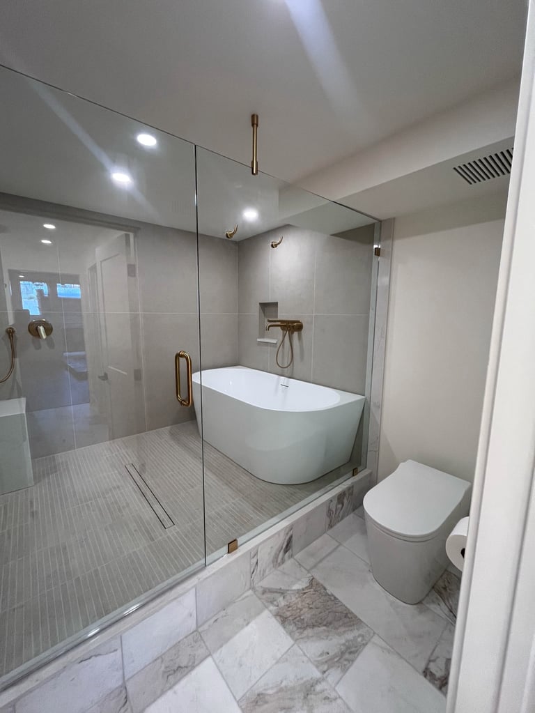 Modern bathroom with white freestanding tub, glass shower enclosure, white toilet, marble and tile flooring, and bronze fixtures