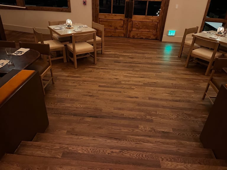 Empty restaurant dining room with wooden tables and chairs on hardwood floor, exit sign visible