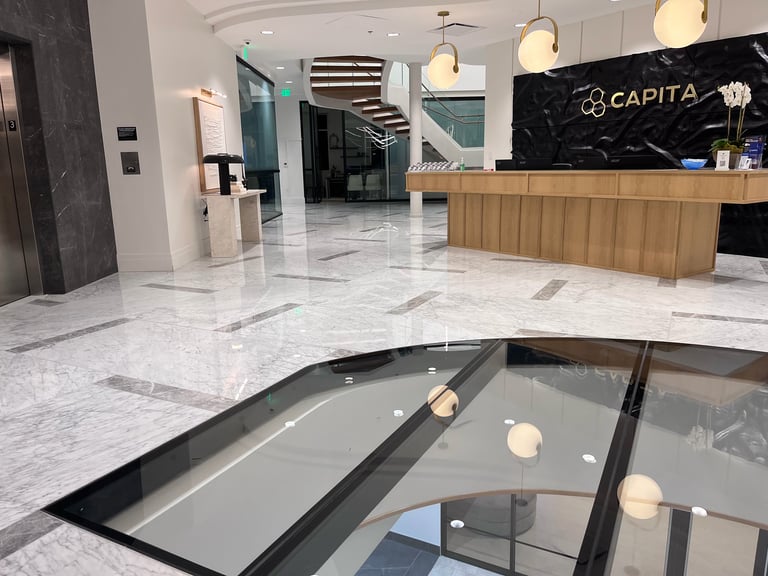 Modern luxury lobby with marble floors, wooden reception desk labeled CAPITA, pendant lights, and a skylight showing upper level